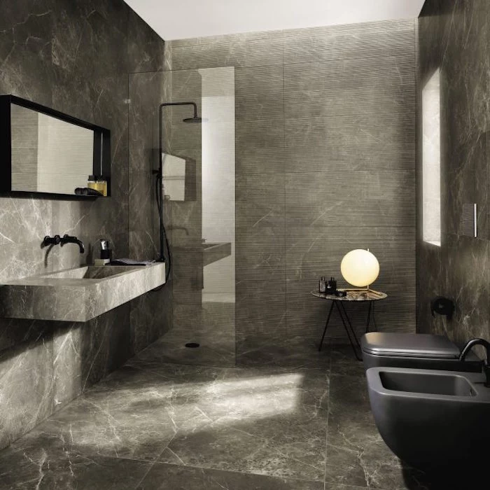 pewter grey room, with stone effect walls and floor, containing a shower, with a clear glass divider, a massive stone sink, with a black faucet, near a mirror in a black frame, dark grey toilet, and matching bidet, modern bathroom ideas