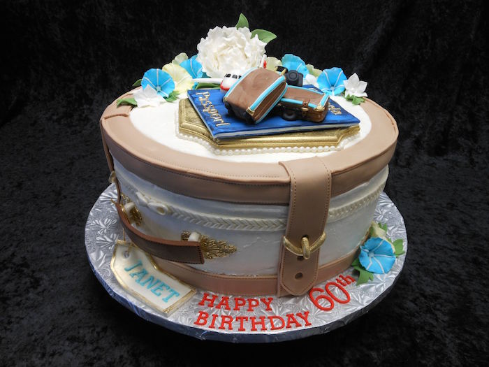 round cake made to look like a suitcase, in white and beige, with golden buckle details, topped with fresh flowers, a passport and small suitcases, made of fondant, happy 60th birthday, written in red frosting