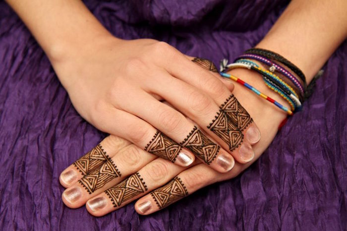 small finger henna tattoos, in dark brown, on a pair of hands, resting one on top of the other, on a crinkled purple fabric