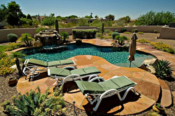 desert plants in a small garden, with pebbles and beige stone tiles, containing a small pool, and three green sun beds