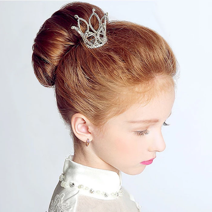 ornament shaped like a small, bejeweled silver crown, decorating the head of a young girl, with auburn hair styled in a bun, white formal dress