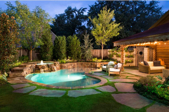 kidney shaped pool, with a small fountain, near a house with a lit porch light, small inground swimming pools, lawn with trees and shrubs