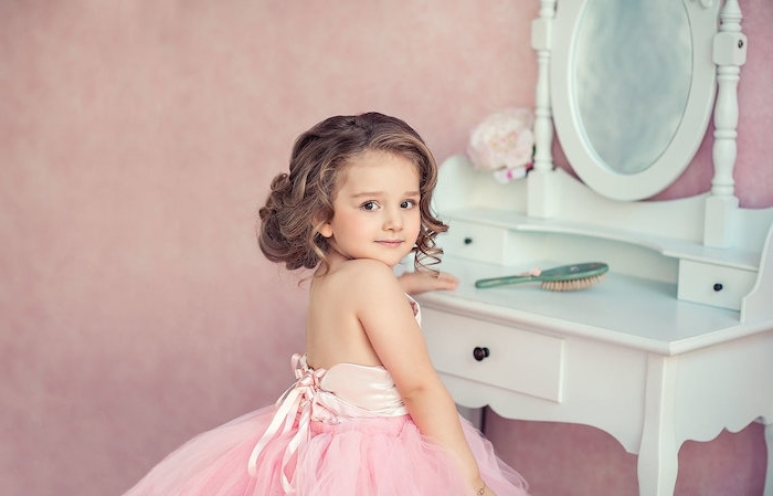 shoulder-length curled brunette hair, worn by a small girl, in a pale pink dress, with a tulle skirt, little girl haircuts, standing in front of a pale blue vanity