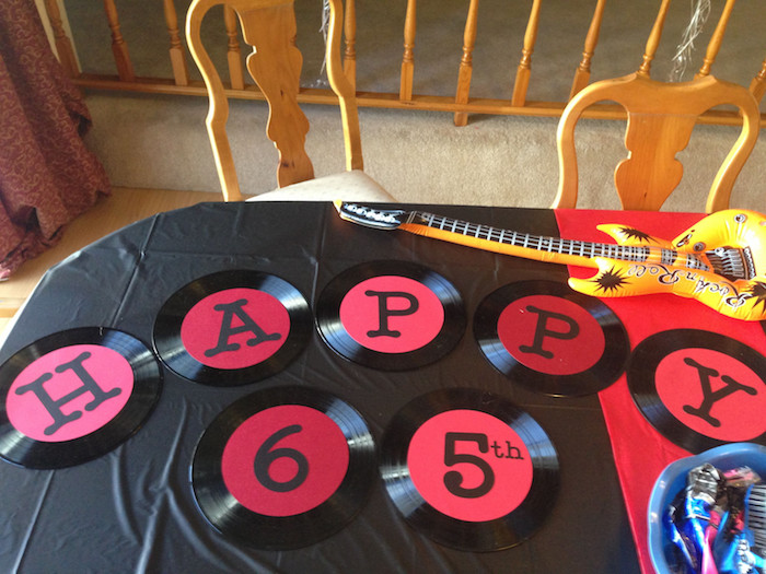 seven vinyl records, in black and red, each decorated with a letter, spelling happy 65th, placed on a table, near an inflatable guitar