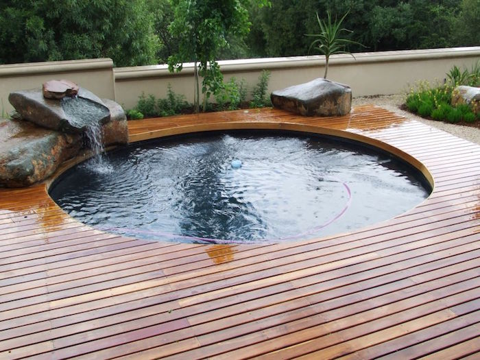 pool patio ideas, round pool surrounded by wooden planks, and decorated with large stones, water flowing into the fool, from one of the stones