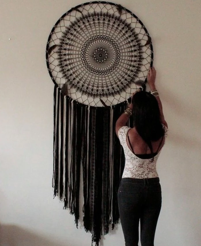 intricate woven pattern in brown, on a large dream catcher, with black tassels, dark-haired woman adjusting it, on a pale cream wall