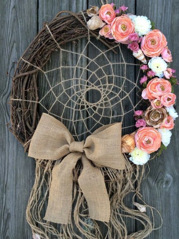 burlap bow and tassels made of plain string, decorating a wreath, adorned with pink and white flowers, several conch shells, and a dreamcatcher net, big dream catchers, grey wooden surface