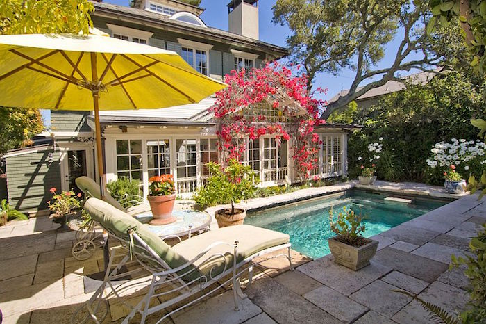 red flowers decorating the entrance of a house, small rectangular pool nearby, two sun beds, and a large yellow umbrella