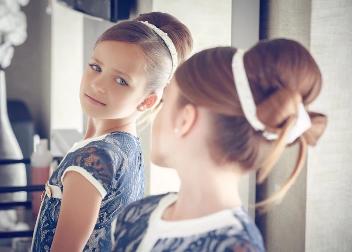 mirror reflection of a young girl, with brunette hair, tied in a bun, and a bow detail, cute girls hairstyles, navy and white lace dress, and a white hairband