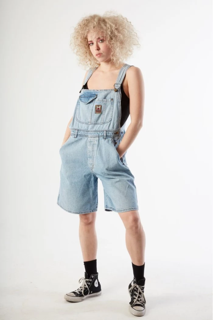 80s clothes, pale knee-length denim overalls, worn by a young woman, with voluminous blonde curly hair
