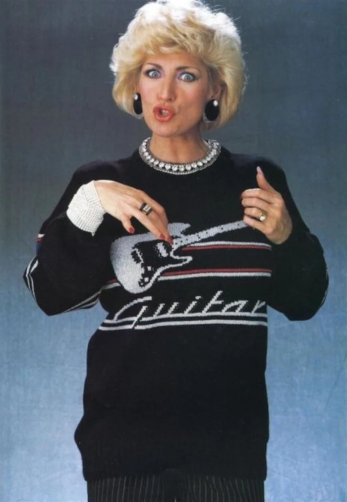 tunic sweater in black, decorated with an image of an electric guitar, and the word guitar, 80's dress up ideas, worn by a woman with short, curly blonde hair, making a funny face