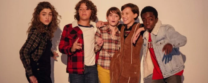 actors from the series stranger things, dressed in typical 80s outfits, plaid jackets and shirts, denim and corduroy