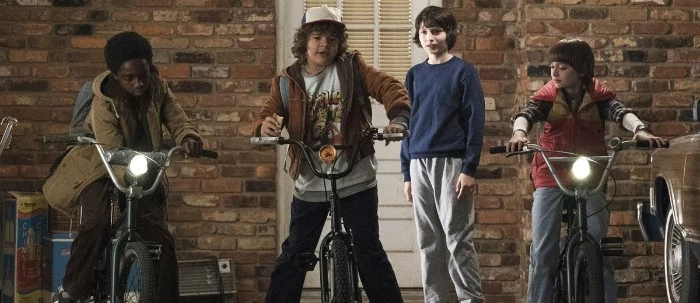 four child actors, from the television series stranger things, dressed in 80s costumes, printed t-shirts and jeans, tracksuit bottoms and corduroy jackets
