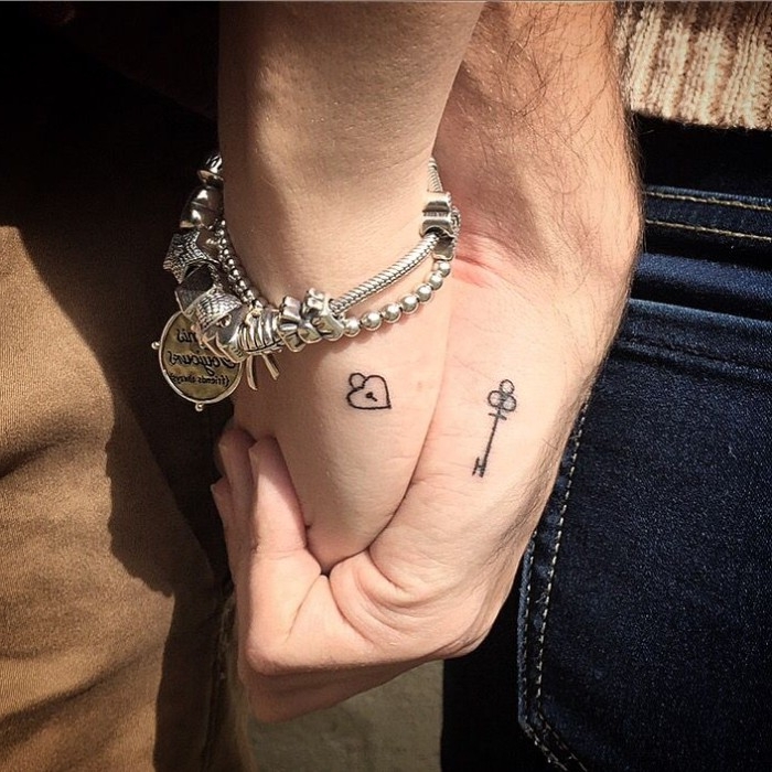 charm bracelet in silver, on a hand with a tiny, heart-shaped padlock tattoo, held by a bigger hand, with a key-shaped tattoo, husband and wife tattoos, with minimalistic designs