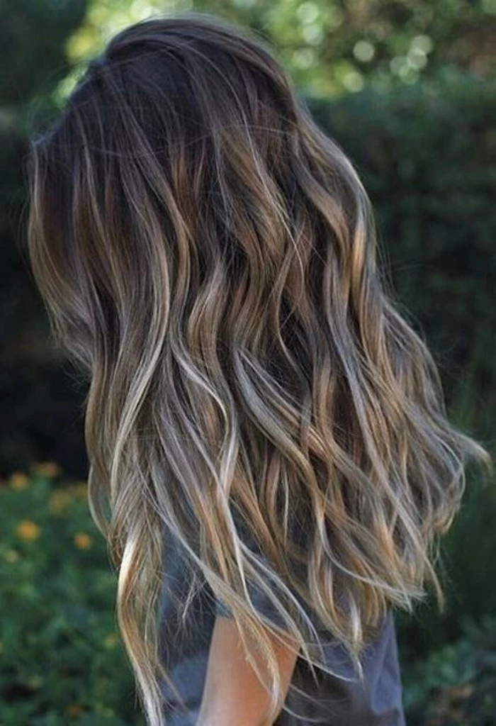balayage hair, long and wavy, with dark roots, and light blonde highlights, on a woman, dressed in a grey t-shirt