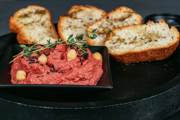 beet hummus recipe, beet hummus in black bowl, garnished with chickpeas and herbs, bruschetta slices on the side