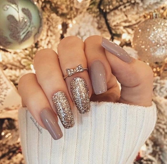 bow-like diamante detail, on a ring, worn by a hand with long, coffin-shaped nails, painted in dark milky grey nail polish, and decorated with gold glitter