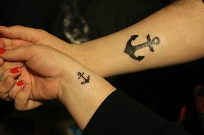 one big and one small anchor, tattooed in black, near the wrists of two linked amrs, one hand has red nail polish