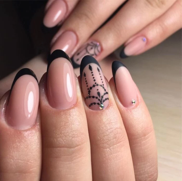 hands with almond shaped nails, in nude pink color, with black tips, and black hand-painted motifs, decorated with rhinestones, some are glossy, while others are matte