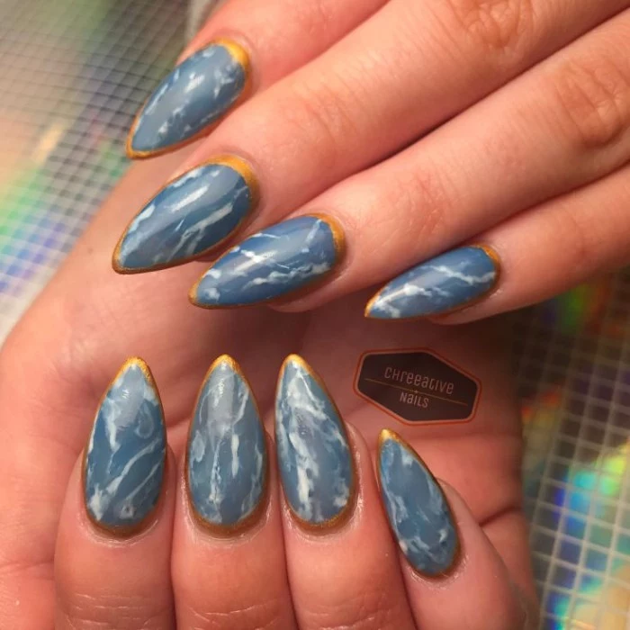 manicure in blue, with a marble-like white pattern, and a gold outline, almond nail designs, on two hands 