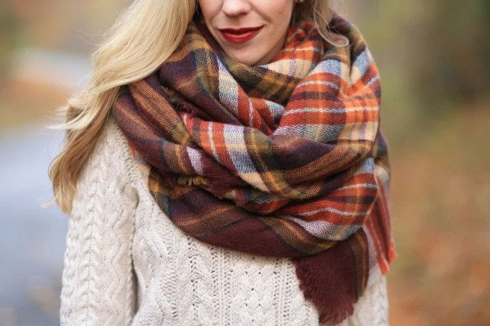 shawl in warm earth tones, wrapped around the neck of a blonde woman, how to tie a blanket scarf, wearing an off-white, cable knit jumper, and dark red lipstick