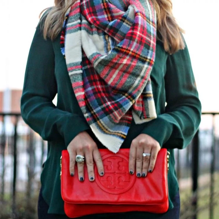 leather bag in red, with a big round embossed logo, held by a woman, in a dark green jumper, accessorized with a plaid blanket shawl, folded around her neck