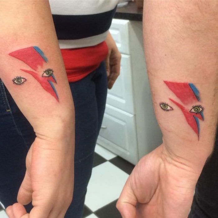 ziggy stardust tattoo, david bowie's eyes, with a red and blue lightning bolt, near the wrists of two arms