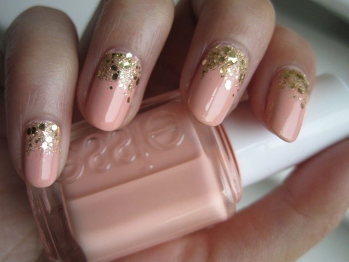 glitter flakes in gold, decorating the base of four oval nails, covered in pink nail polish, nude nail designs, on a hand, holding a nail polish bottle