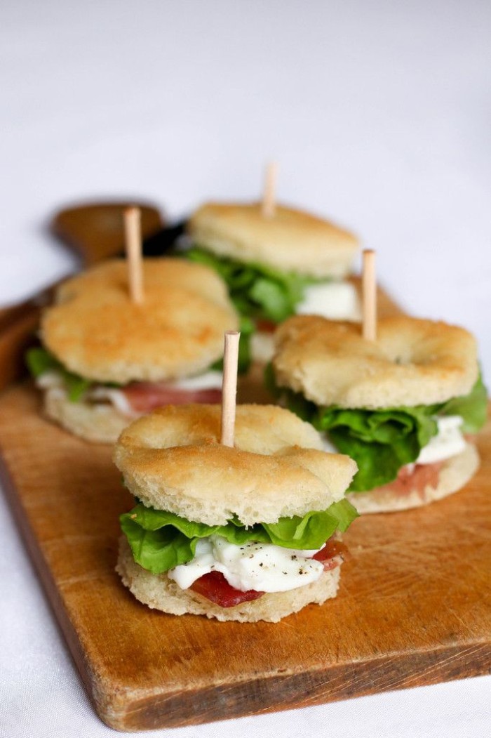 round party sized sandwiches, containing prosciutto and mozzarella, with fresh lettuce, hor dourves on a chopping board, made of wood
