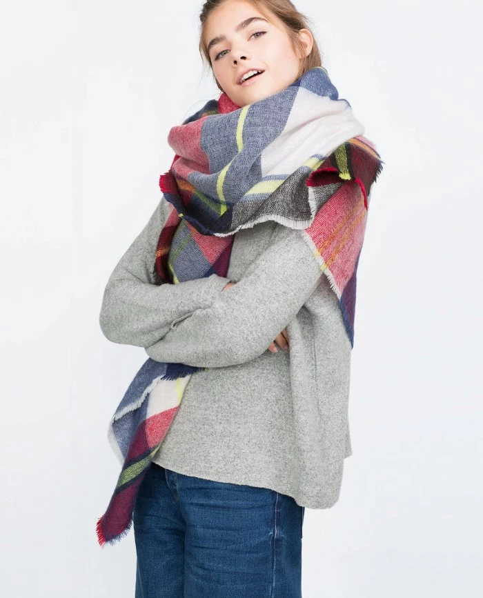 how to wear a blanket scarf, smiling young woman, with arms crossed, wearing a pale grey jumper, dark blue jeans, and an oversized scarf, around her neck