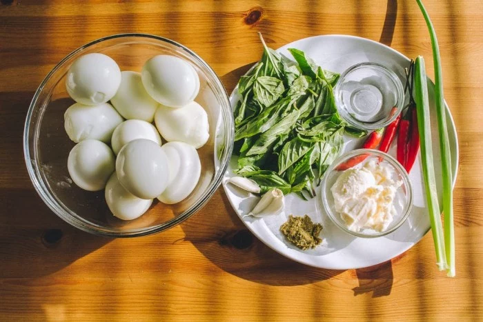 glass bowl filled with hardboiled eggs, next to a white plate, containing ingredients for horderves, mayo and garlic cloves, spring onion and chilis, basil and pesto