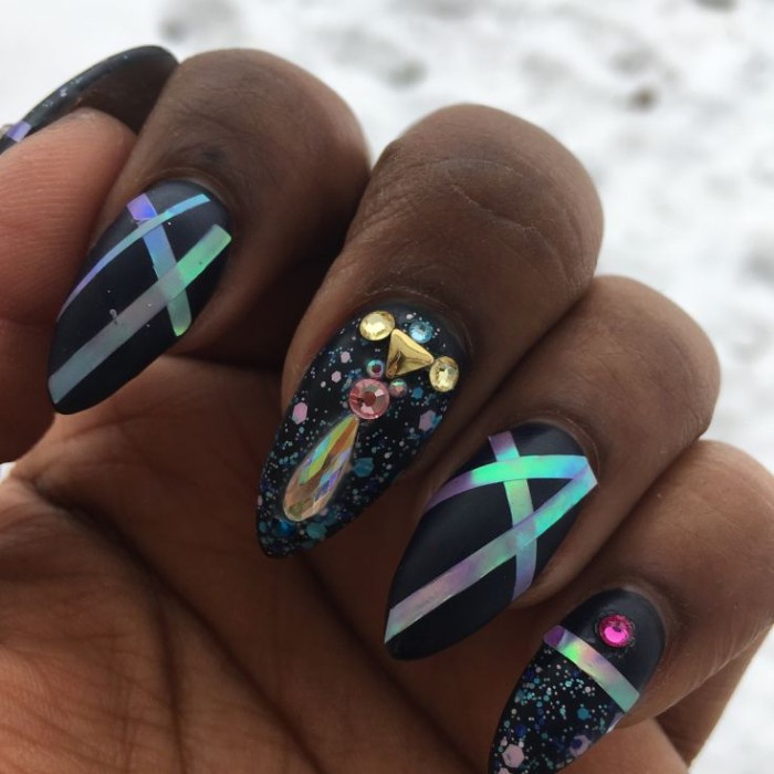 holographic stripes and iridescent glitter flakes, on ablack acrylic nail shapes, decorated with rhinestones, on a black hand