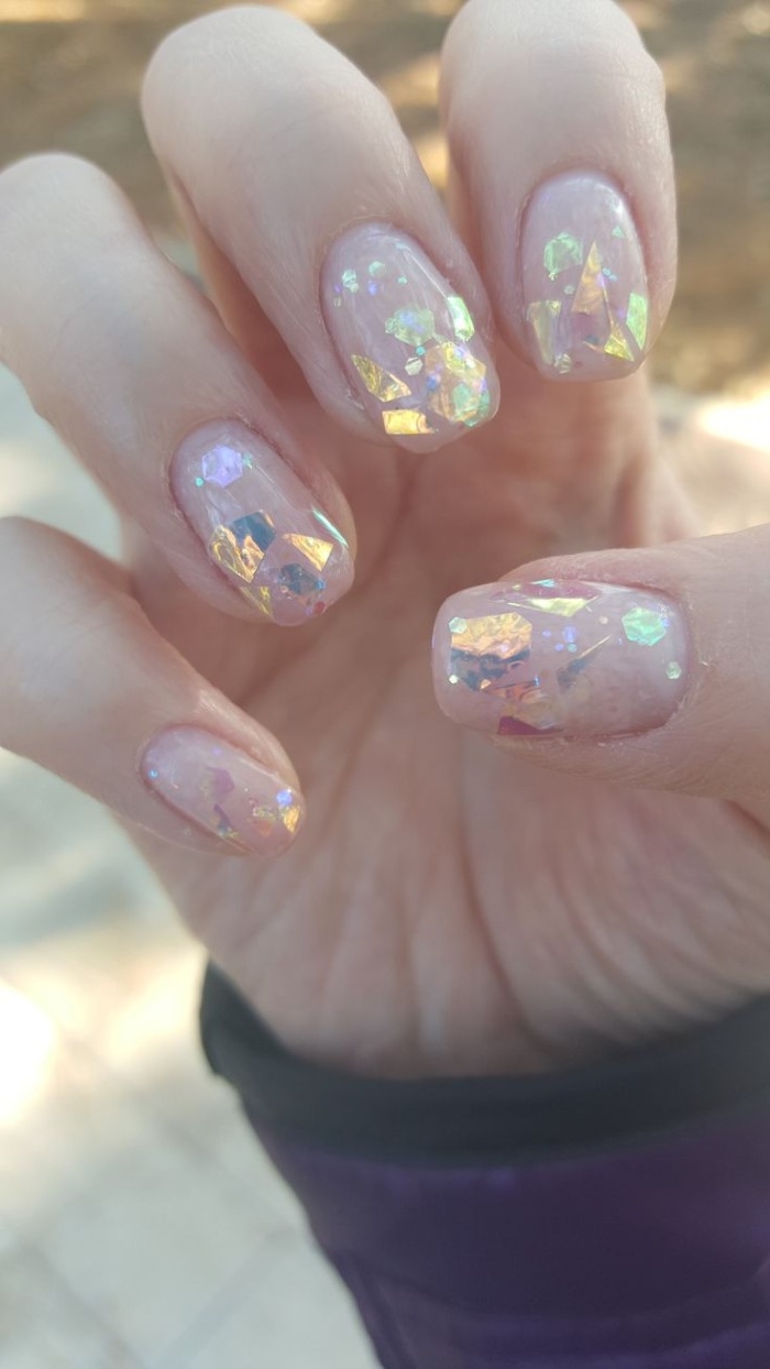 large flakes of iridescent glitter, on the square manicure of a hand, with folded fingers, and nude pink nail polish