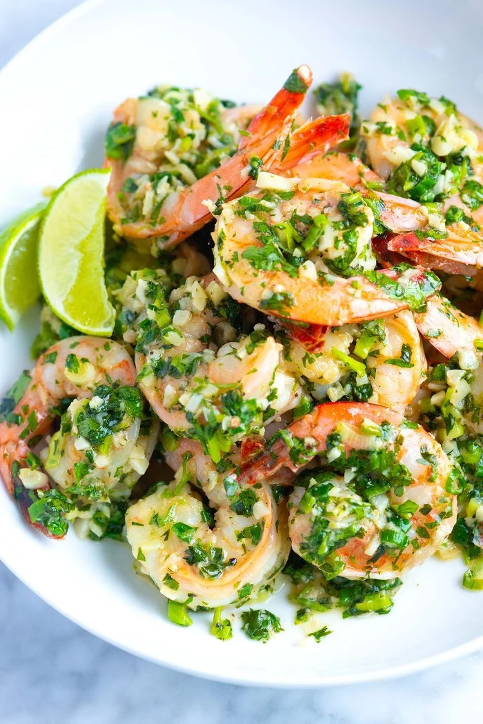 seasoned jumpo prawns, with chopped fresh green herbs, hors dourves, inside a white plate, with lemon and lime wedges