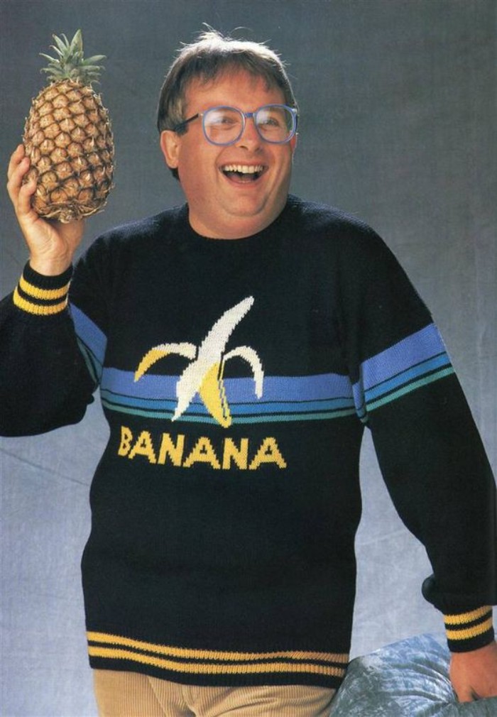sweater in black, yellow and blue, with a picture of a banana, and the word banana in yellow, 80s outfits guys, worn by a laughing man, with glasses, holding a pineapple