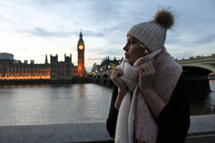 big ben in the evening, near a woman, dressed in winter clothing, knitted beanie hat, with a fur pom pom, large soft scarf around her neck, how to fold a blanket scarf 