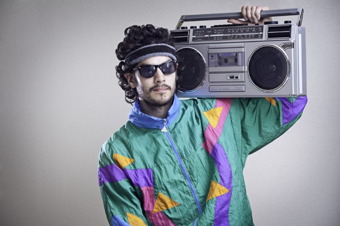 teal jacket with yellow and light blue, pink and purple geometric shapes, worn by a man with dark curly hair, a headband and sunglasses, 80s clothes, holding a boombox