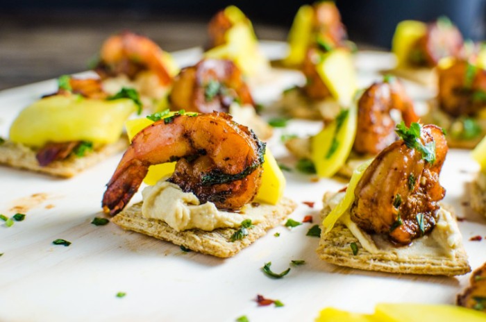 square crackers with hummus, topped with mango, and grilled prawns, hour derves sprinkled with fresh, chopped green herbs