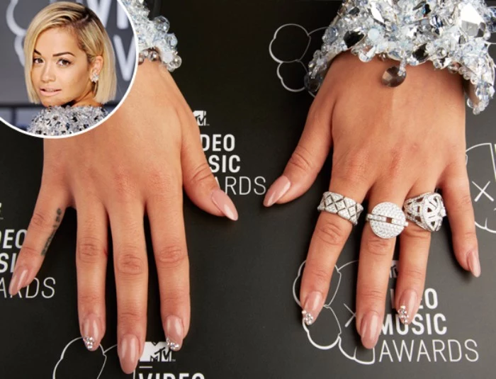 rita ora's manicure, nude pink oval nails, decorated with rhinestones, on hands decorated with chunky silver rings, and big bracelets