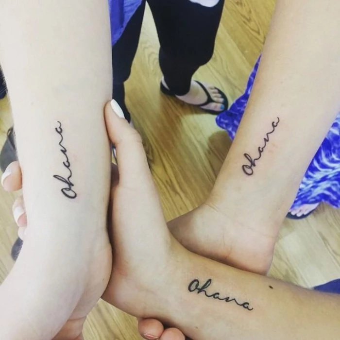three linked hands, featuring identical black tattoos, of the word ohana, near their wrists, matching sister tattoos, also suitable for cousins 