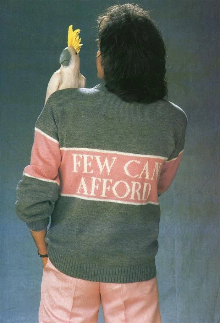 view from the back of a man, with medium length black wavy hair, holding a parrot doll on his shoulder, and dressed in a grey, pink and white sweater, with an inscription saying, few can afford, 80s outfits guys