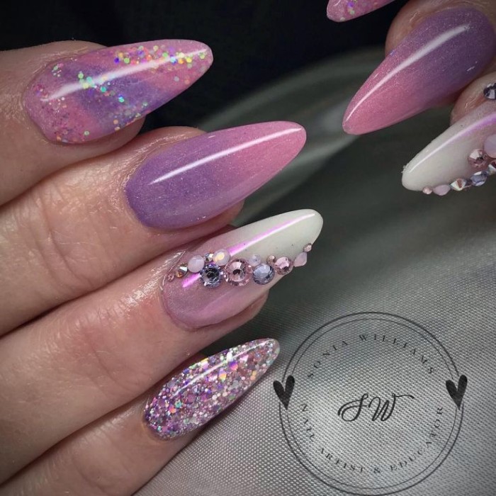 violet and pink nail polish, decorated with glitter, and iridescent rhinestones, on a long and sharp amnicure, seen in close up