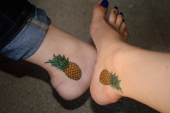 orange and green tattoos, done between the ankles and heels of two feet, and depicting two identical pineapples, matching sister tattoos, one foot has dark blue nail polish, and a toe ring