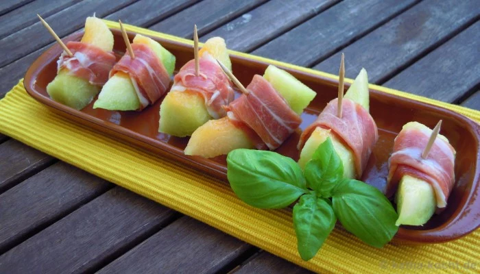 melon pieces wrapped in prosciutto, hor dorves, inside a brown oval plate, decorated with a sprig of fresh basil