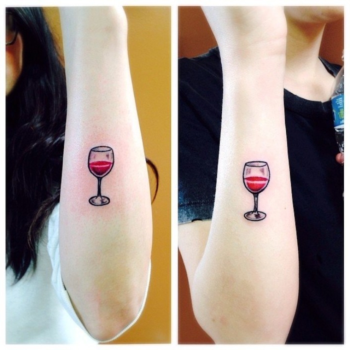 glasses half-filled with red wine, identical tattos for firends, done with red and black ink, on the lower part, of two pale arms