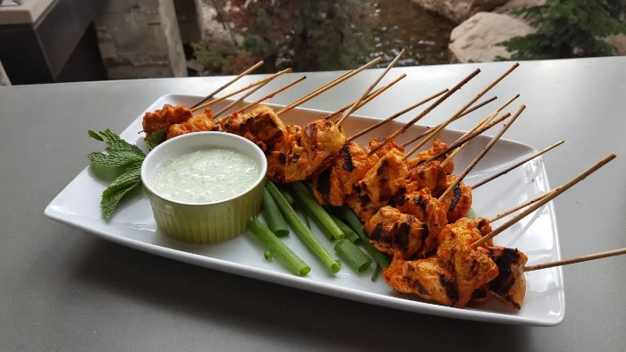 hour derves, grilled chicken skewers, on a white rectangular plate, near spring onions, fresh mint leaves, and a bowl with yoghurt dip