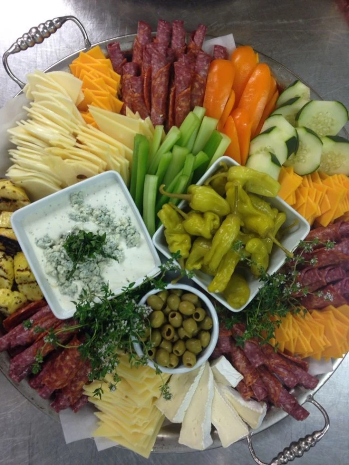 metal tray with a selection of hor dorves, different kinds of cheese and salami, jalapenos and pitted olives, chopped vegetables and yoghurt dip