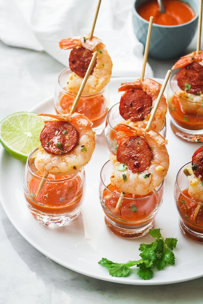 shot glasses containing an orange sauce, and skewers with prawns and chorizo, hor dourves on a white round plate