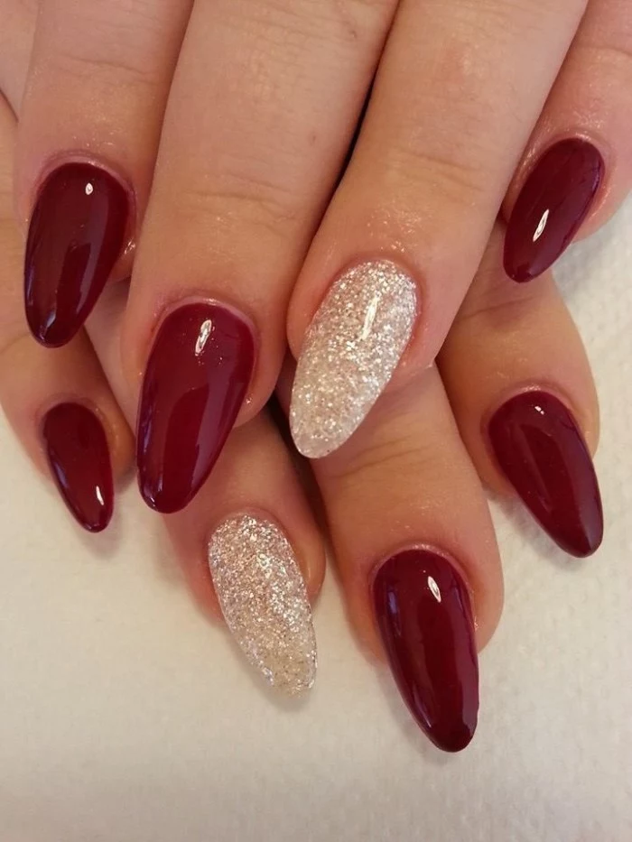 classic red nail polish, worn on two pale hands, with oval shaped nails, resting on a white surface, the ring finger nails are decorated with fine silver glitter
