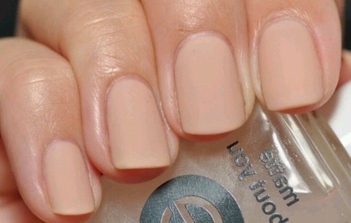 manicure with short square nails, painted in a nude pink nail polish, seen in close up, on a hand holding a clear bottle of nail polish